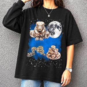 Könnte beinhalten: Schwarzes übergroßes T-Shirt mit einem grafischen Design von drei Schlangen, einem Vollmond und einem Sternenhimmel. Die Schlangen sind braun und beige auf blauem Hintergrund.