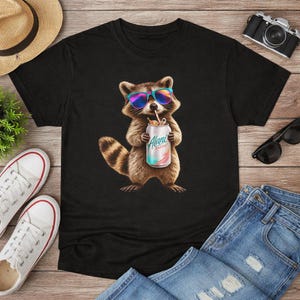 May include: Black t-shirt featuring a raccoon wearing sunglasses and holding a can of "Alani Energy". The raccoon is brown and white with a striped tail. The can is white with a colorful design. The shirt is surrounded by accessories.