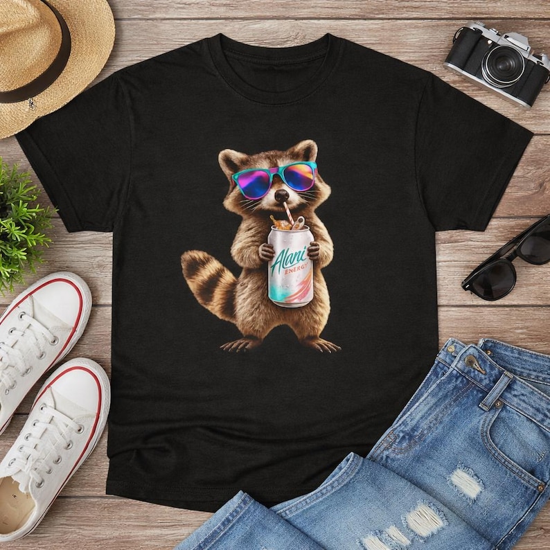 May include: Black t-shirt featuring a raccoon wearing sunglasses and holding a can of "Alani Energy". The raccoon is brown and white with a striped tail. The can is white with a colorful design. The shirt is surrounded by accessories.