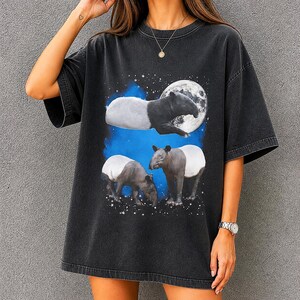 Puede incluir: Camiseta negra con un diseño de tres tapires, una luna llena y un cielo estrellado. Los tapires son en blanco y negro con un acento azul y blanco. La camiseta tiene mangas cortas y un corte holgado.