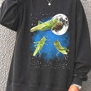May include: Dark gray sweatshirt featuring a graphic of green grasshoppers against a blue and starry background with a full moon. The sweatshirt has long sleeves and a crew neck.