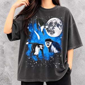 May include: Dark gray t-shirt featuring a graphic of three colobus monkeys against a blue and starry background with a full moon. The monkeys are black and white with long tails. The shirt has short sleeves and a relaxed fit.