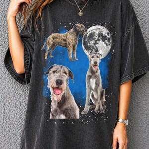 May include: Dark grey t-shirt with a graphic of three dogs, a full moon, and stars. The dogs are different breeds and poses. The background is a blue hue with white stars. The shirt is a dark grey colour.