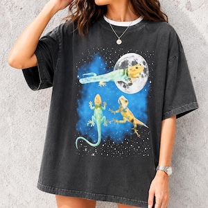 May include: A charcoal grey short-sleeve t-shirt featuring a graphic print of three geckos against a blue nebula background with a full moon. The geckos are depicted in shades of yellow, green, and blue, surrounded by small white stars.