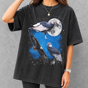 Op de afbeelding: Een antracietzwart t-shirt met korte mouwen en een grafische print van twee Andescondors en een valk tegen een blauwe sterrenhemel met een grote volle maan. De vogels zijn afgebeeld in vlucht en zittend op een ondergrond.