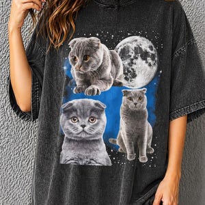 K&ouml;nnte beinhalten: Dunkelgraues T-Shirt mit einer Grafik von drei grauen Scottish Fold-Katzen und einem Vollmond vor einem blauen und sternen&uuml;bers&auml;ten Hintergrund. Die Katzen haben gelbe Augen und sind in verschiedenen Posen. Das Shirt hat kurze &Auml;rmel.