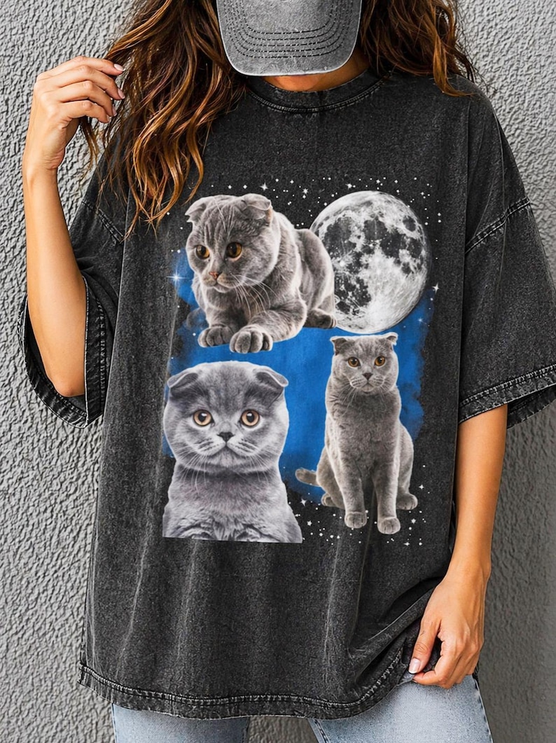 K&ouml;nnte beinhalten: Dunkelgraues T-Shirt mit einer Grafik von drei grauen Scottish Fold-Katzen und einem Vollmond vor einem blauen und sternen&uuml;bers&auml;ten Hintergrund. Die Katzen haben gelbe Augen und sind in verschiedenen Posen. Das Shirt hat kurze &Auml;rmel.