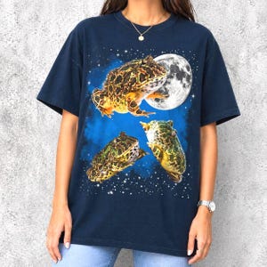 Può includere: T-shirt blu navy con una grafica di tre rane, una luna piena e un cielo stellato. Le rane sono marroni, verdi e gialle. La luna è bianca e le stelle sono piccoli punti bianchi.