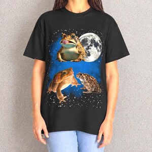 May include: Black t-shirt featuring a whimsical design of three frogs against a blue and starry background, with a full moon. The top frog has its tongue out. The frogs are in shades of brown and tan.