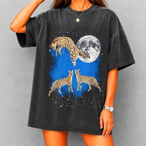 Puede incluir: Camiseta gris oscuro con un gráfico de un leopardo saltando frente a una luna llena y estrellas. Dos leopardos están de pie debajo. El diseño tiene un fondo de nube azul.
