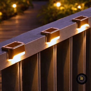 May include: Three bronze-colored solar-powered lights are mounted on a gray railing. Each light has a rectangular top and a downward-facing light source, casting a warm glow. The background features a blurred view of a wooden walkway and foliage.