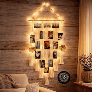 May include: A wall-mounted photo display with fairy lights. The display features a wooden dowel and hanging strings with small photos, tassels, and decorative leaves. The warm glow of the lights illuminates the photos and the wooden wall behind it.
