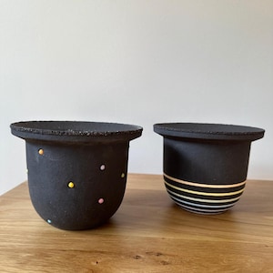 Puede incluir: Dos macetas de cerámica negra. Una tiene lunares de colores, la otra rayas horizontales en colores pastel. Ambas tienen un borde texturizado y una base redondeada. Están sobre una superficie de madera.