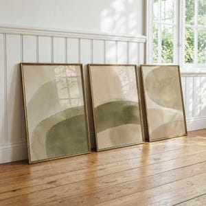 Könnte beinhalten: Drei rechteckige Wandbilder in dünnen Goldrahmen mit abstrakten Pinselstrichen im Aquarellstil. Die Kunstwerke zeigen geschichtete, geschwungene Formen in verschiedenen Schattierungen von Salbeigrün, Olivgrün und Creme mit weichen, fließenden Übergängen.