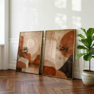 May include: Two rectangular framed wall art prints featuring abstract landscapes in shades of terracotta, beige, and dark brown. The artwork displays geometric shapes, circular sun-like forms, and thin, dark, spindly tree silhouettes against a textured background.
