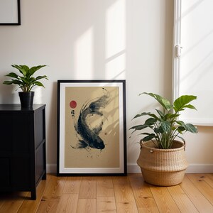May include: Framed art print featuring a black and white koi fish illustration with a red sun and Japanese characters. The artwork is displayed on a light tan background, framed in black, and leaning against a white wall. A black cabinet and potted plants are in the foreground.