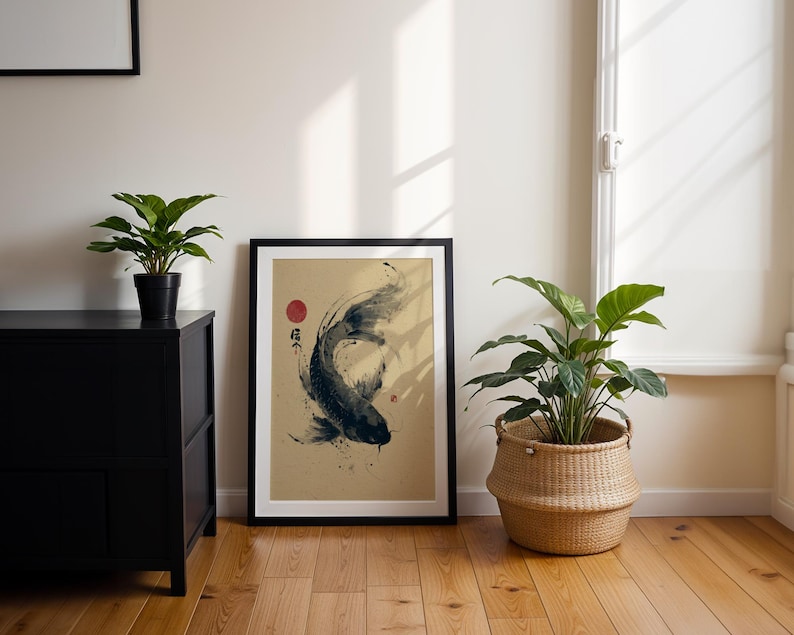May include: Framed art print featuring a black and white koi fish illustration with a red sun and Japanese characters. The artwork is displayed on a light tan background, framed in black, and leaning against a white wall. A black cabinet and potted plants are in the foreground.