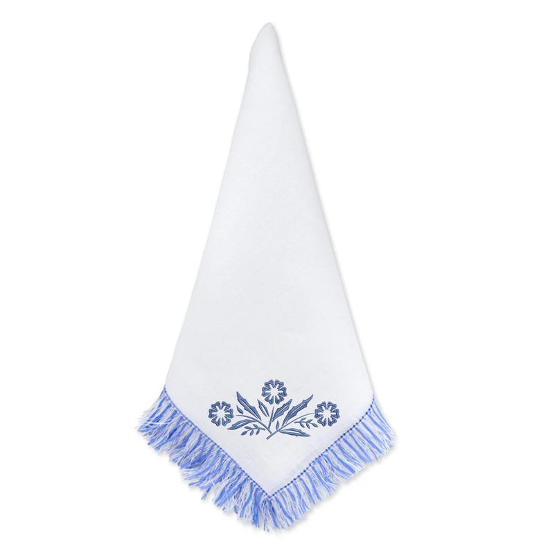 May include: White linen napkin with a blue floral embroidery design and fringed edges. The embroidered design features three stylized flowers and leaves. The napkin is folded into a triangle shape.