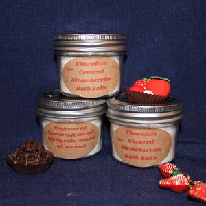 May include: Three glass jars with silver lids, labelled "Chocolate Covered Strawberries Bath Salts." One jar has a strawberry decoration. Ingredients include fragrance oil, Epsom salt, baking soda, and coconut oil.