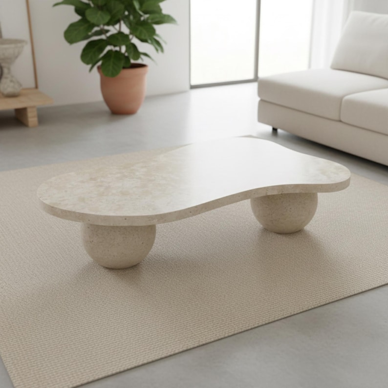 May include: A beige, organically shaped coffee table with a speckled surface, supported by two rounded, pedestal-style legs. The table sits on a light beige rug in a modern living room setting.