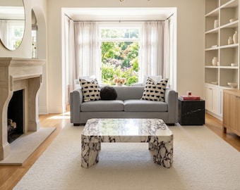 May include: A living room with a gray sofa, patterned pillows, and a marble coffee table. The room features a fireplace, built-in shelves, and a large window with sheer curtains. A white rug covers the floor.