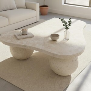 May include: A cream-colored, organically shaped coffee table with two rounded pedestal bases. The table surface holds a small bowl, a book, a mug, and a glass vase with greenery. The table sits on a light beige rug.