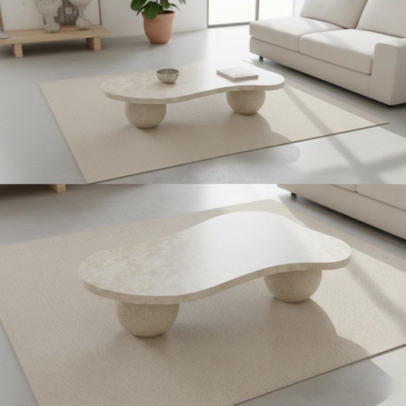 May include: A modern, beige coffee table with a unique, organic shape. The tabletop is supported by two rounded, spherical bases. The table sits on a neutral-toned rug, creating a minimalist aesthetic. The table's dimensions are approximately 47 inches long and 24 inches wide.