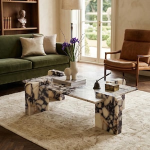 May include: A marble coffee table with a unique, geometric design. The table features a white and dark purple veined pattern. A green velvet sofa, a brown leather chair, and a beige rug are also visible in the room.