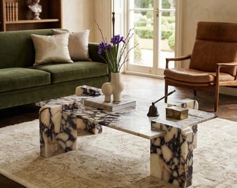May include: A marble coffee table with a unique, geometric design. The table features a white and dark purple veined pattern. A green velvet sofa, a brown leather chair, and a beige rug are also visible in the room.