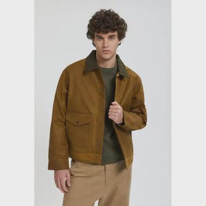 Puede incluir: Chaqueta marrón con cuello verde y dos bolsillos delanteros. La chaqueta se lleva sobre una camisa verde y se combina con pantalones color beige. El modelo está de pie frente a un fondo blanco.