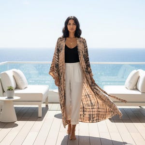 May include: A woman wearing a long, flowing tie-dye kimono in shades of tan and black over a black top and white pants. The kimono has wide sleeves and a matching tie belt. The background features a blue ocean and a sunny sky.