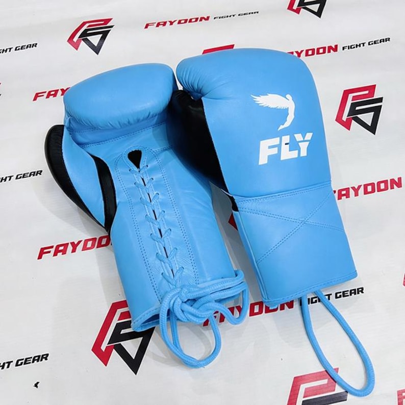 May include: A pair of light blue boxing gloves with black detailing. The gloves feature a lace-up design and the word "FLY" is printed on the wrist area. These gloves are designed for fight gear.