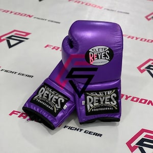 以下が含まれることがあります： 黒と白の「CLETD REYES」ロゴが入った紫色のボクシンググローブのペア。 グローブは光沢のある素材でできており、プロの使用のために設計されています。 グローブは、「FIGHT GEAR」と「FRYOON」の文字が印刷された白い表面にあります。
