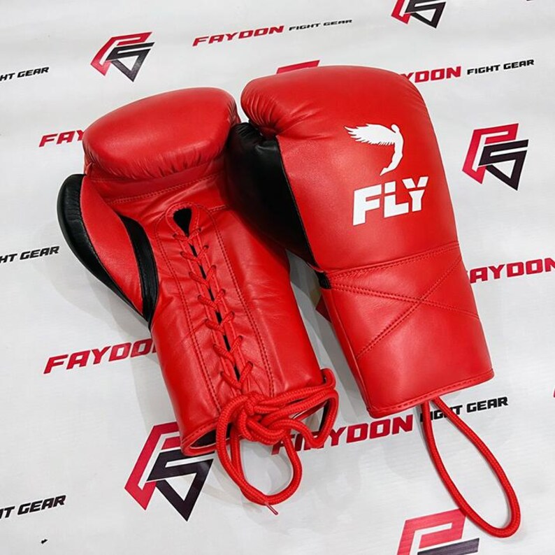 May include: Two red boxing gloves with black detailing. The gloves feature the word "FLY" and a bird graphic. They are laced with red laces. The background has the text "FAYDON FIGHT GEAR" printed on it.