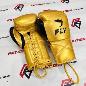 May include: A pair of gold boxing gloves with black detailing. The gloves feature a lace-up closure and the word "FLY" is printed in black on one glove. These gloves are designed for combat sports.