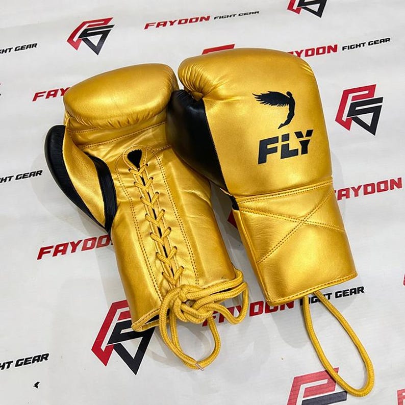 May include: A pair of gold boxing gloves with black detailing. The gloves feature a lace-up closure and the word "FLY" is printed in black on one glove. These gloves are designed for combat sports.