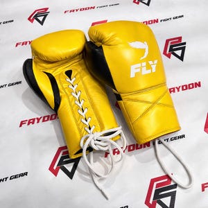 May include: A pair of yellow boxing gloves with black detailing. The gloves feature white laces and the word "FLY" is printed on one glove. The background has the words "FAYDON FIGHT GEAR" in red.