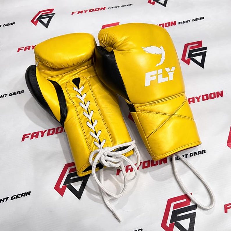 May include: A pair of yellow boxing gloves with black detailing. The gloves feature white laces and the word "FLY" is printed on one glove. The background has the words "FAYDON FIGHT GEAR" in red.