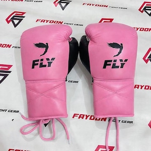 May include: A pair of pink boxing gloves with black detailing. The gloves display the word "FLY" in black, along with a black wing design. Pink laces are visible at the wrist.