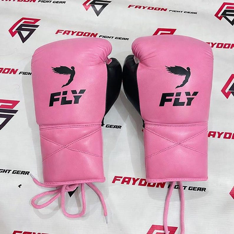 May include: A pair of pink boxing gloves with black detailing. The gloves display the word "FLY" in black, along with a black wing design. Pink laces are visible at the wrist.