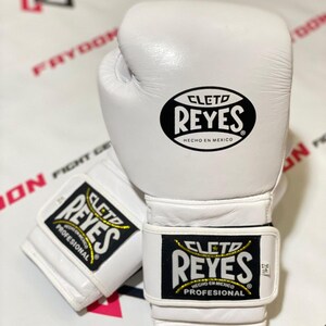 以下が含まれることがあります： 黒と金の「CLETO REYES」ロゴが入った白い革製ボクシンググローブ。グローブには「HECHO EN MEXICO」と「PROFESIONAL」のラベルが付いています。片方のグローブは手首のストラップが開いており、もう片方は閉じています。