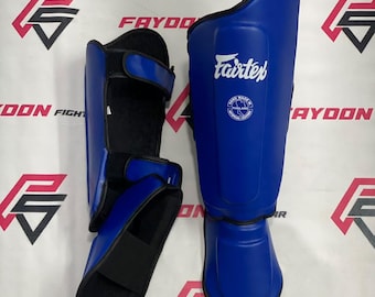 Op de afbeelding: Een paar blauwe scheenbeschermers met het witte Fairtex-logo en een rond embleem met de tekst Hand Made In Thailand Artificial Leather. De beschermers hebben een zwarte voering, zwarte randen en verstelbare klittenbandsluitingen.