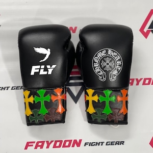 Puede incluir: Un par de guantes de boxeo negros con logotipos blancos. El guante izquierdo muestra una figura alada y el texto FLY. El guante derecho presenta un emblema circular de CHROME HEARTS. Ambos puños están decorados con parches de cuero en forma de cruz.