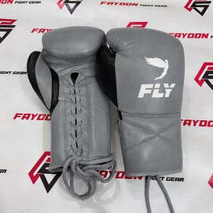 May include: A pair of grey boxing gloves with black detailing. The gloves feature a lace-up design and the word "FLY" is printed in white, along with a white graphic of a bird. The gloves are set against a background with the text "FAYOON FIGHT GEAR".