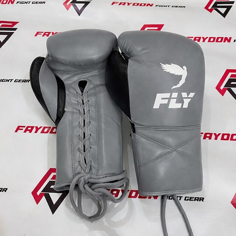 May include: A pair of grey boxing gloves with black detailing. The gloves feature a lace-up design and the word "FLY" is printed in white, along with a white graphic of a bird. The gloves are set against a background with the text "FAYOON FIGHT GEAR".