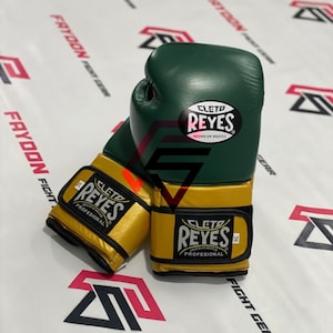Peut inclure: Une paire de gants de boxe verts et dorés avec la marque "CLETD REYES" visible. Les gants sont en cuir et ont un design professionnel. Les gants sont sur une surface blanche avec des motifs graphiques rouges et noirs.
