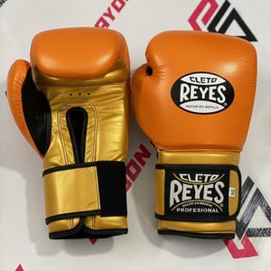 May include: A pair of orange and gold boxing gloves. The gloves have a gold palm and wrist area, with a black strap. The brand name "CLETTO REYES" is visible on the gloves.