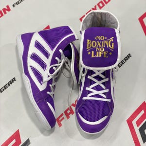 Puede incluir: Un par de botas de boxeo de caña alta en color morado y blanco. Las botas cuentan con paneles de malla, cordones blancos y una lengüeta prominente con el texto dorado NO BOXING NO LIFE impreso en el frente. Diseño con superposiciones blancas.