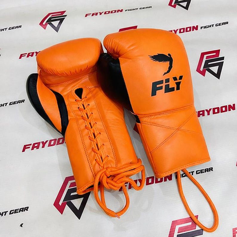 May include: A pair of orange boxing gloves with black detailing. The gloves feature the word "FLY" and a graphic. They are laced with orange laces. The gloves are designed for combat sports.