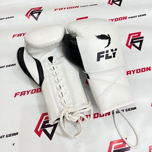 May include: A pair of white boxing gloves with black detailing. The gloves feature a lace-up closure and the word "FLY" is printed on one glove, along with a bird graphic. These gloves are designed for combat sports.